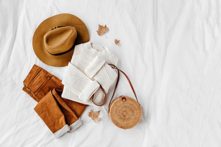 White knitted woolen sweater and bamboo bag with brown trousers and hat on white bed.