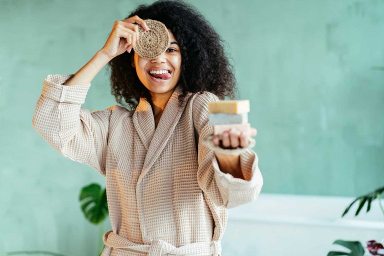 Woman with personal hygiene items holding three soap, covering eye with eco-friendly and zero-waste natural handmade jute washcloth is seen in modern interior bathroom.